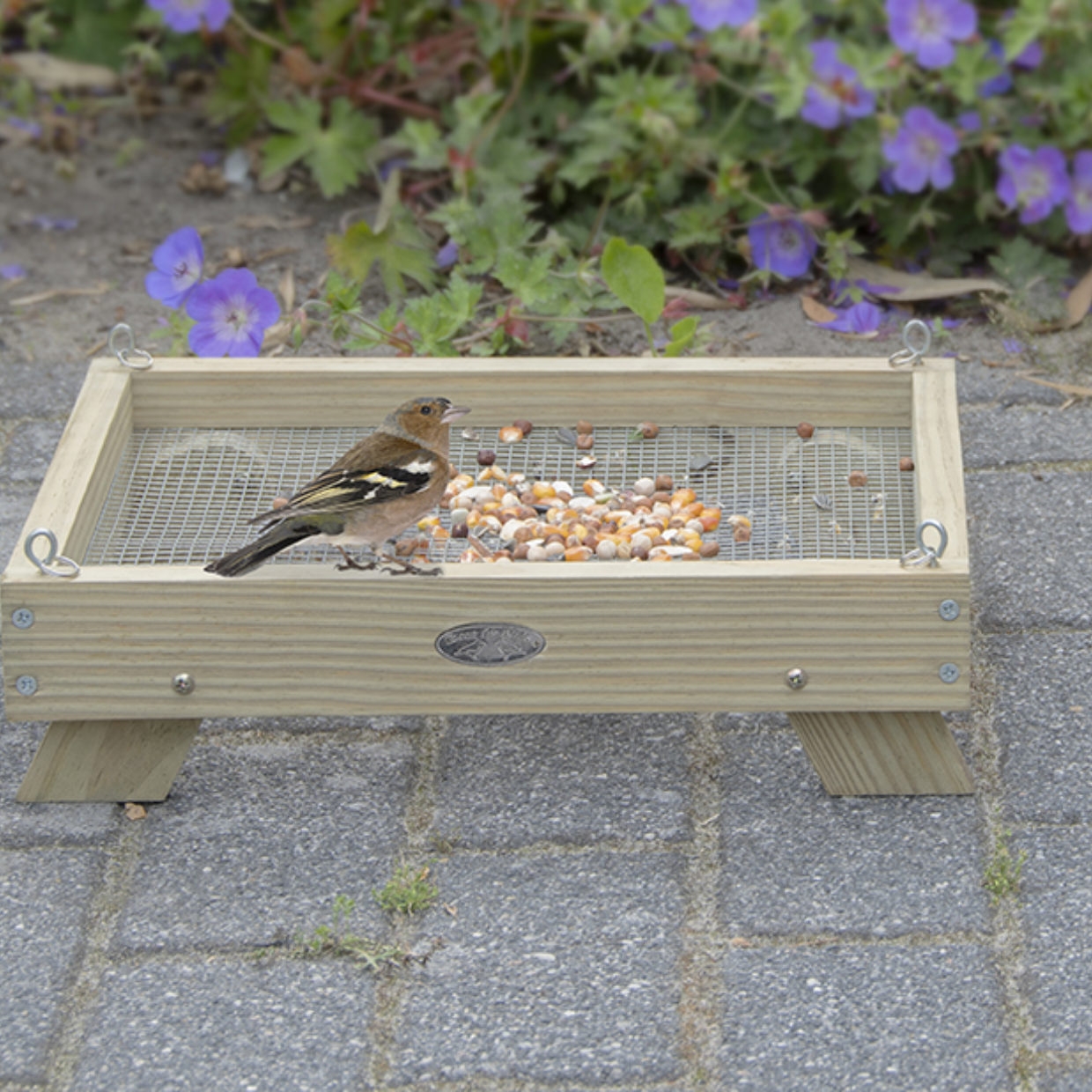 Een houten vogelvoedertafel met een vink die zaden eet. De voedertafel staat op poten op een stenen terras, met bloemen op de achtergrond.